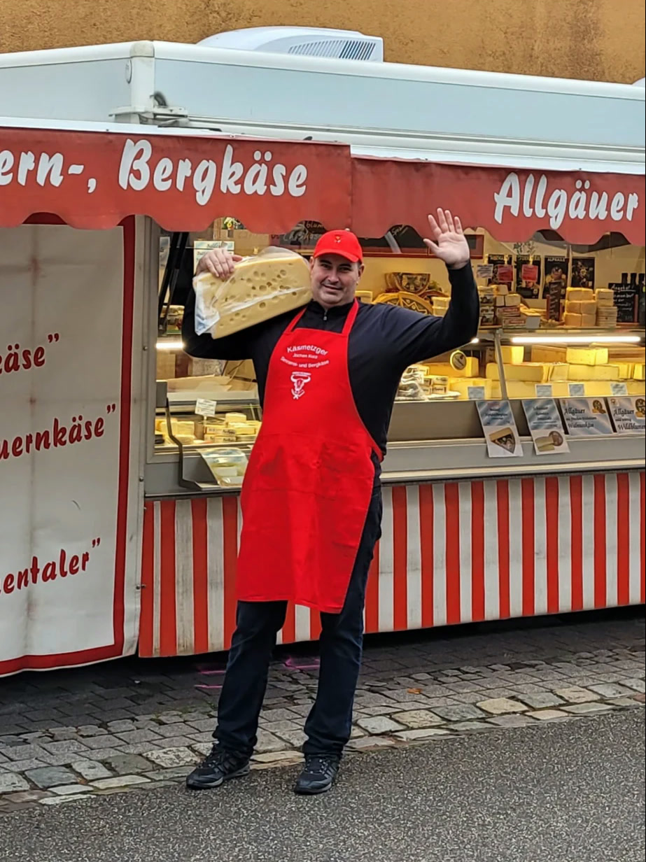 Jochen Kurz trägt ein großes Käsestück vor dem Käsewagen und winkt